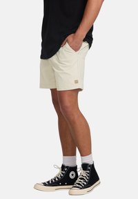 Beige shorts with side pockets, silver hardware, and a logo patch. Paired with black high-top sneakers and white socks.