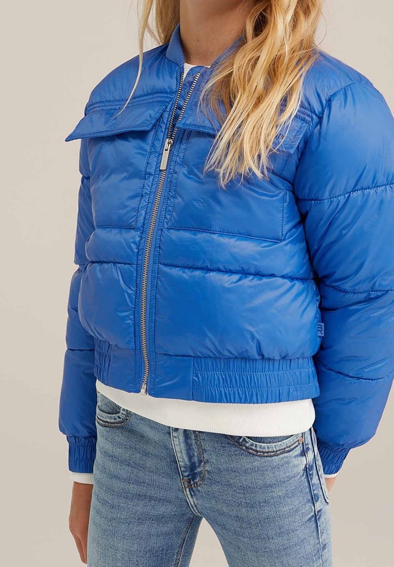Femme portant une veste matelassée bleu vif avec une fermeture éclair à l'avant, des poignets et un ourlet côtelés, associée à un jean bleu clair et un haut blanc.