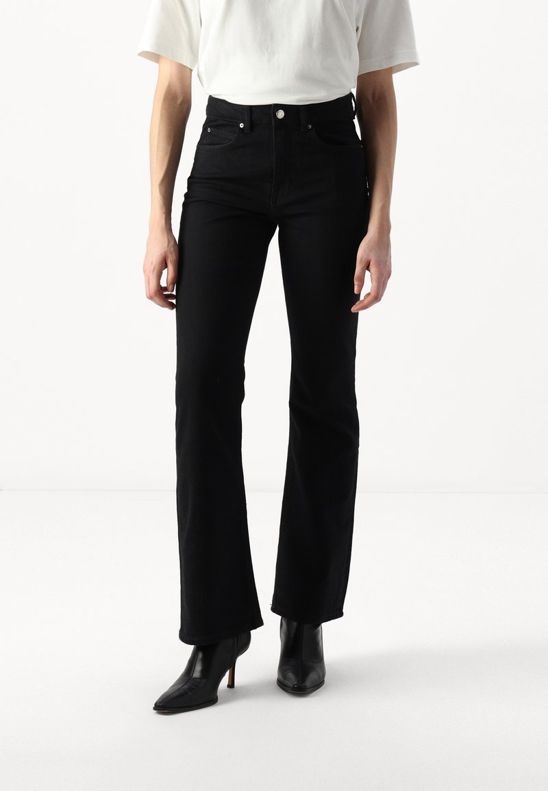 Jeans pretos de perna larga em denim, com cintura alta, bolsos frontais e acessórios prateados. Combinados com botas de cano curto justas pretas com salto.