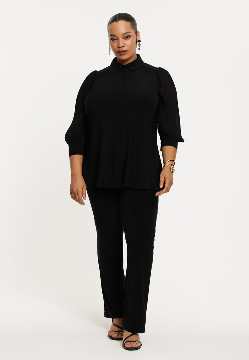 Schwarze Bluse mit Knöpfen und Puffärmeln, kombiniert mit schwarzen geraden Hosen. Glattes Material, minimalistisches Design mit subtilen Details.