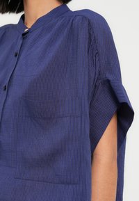 Chemise boutonnée en coton bleu foncé avec des rayures verticales subtiles, manches courtes, col mao et une poche sur le côté gauche.