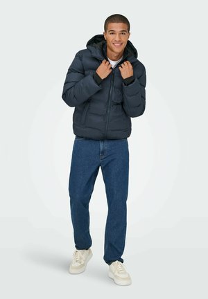 Giubbotto imbottito di un blu navy scuro, con chiusura a zip, tasche frontali e cappuccio, abbinato a jeans blu e sneakers bianche. Tessuto liscio, design trapuntato.