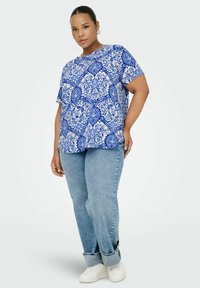 Kvinna som står klädd i en kortärmad skjorta med blå och vita mönster, ljusblå jeans med uppvikta ben, vita sneakers och silverfärgade ringörhängen.