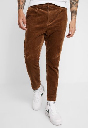 Mann trägt braune Cordhosen und weiße Nike Air-Sneaker, steht mit sichtbaren tätowierten Unterarmen.