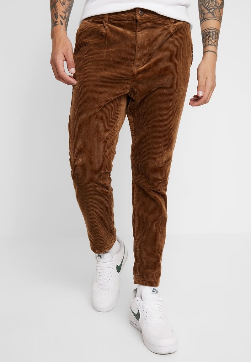 Homme portant un pantalon en velours côtelé marron et des baskets blanches Nike Air, debout avec les avant-bras tatoués visibles.