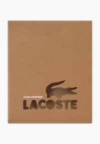 Smeđa kartonska ambalaža proizvoda s izrezanim logotipom krokodila i bijelim natpisom "MON PREMIER LACOSTE."