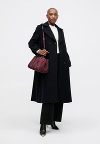 Persona che indossa un lungo cappotto nero, un maglione bianco, pantaloni neri, stivali col tacco neri e tiene in mano una borsa a tracolla di un rosso scuro.