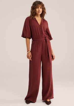 Ženska, oblečena v širok hlačni kombinezon rjave barve s puhastimi rokavi, gumbi spredaj in pasom, ki je zavezan, stoji pred enobarvnim ozadjem.