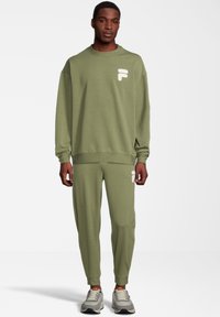 Conjunto de sudadera y pantalones jogger en verde oliva de tejido suave. Presenta un logo blanco texturizado en el pecho y en el bolsillo. Corte ajustado con puños acanalados.