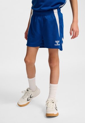 Persoon die blauwe sportkorte broek met witte zijstrepen draagt, witte sokken tot aan de enkels en witte sneakers met zwarte chevronlogo's.