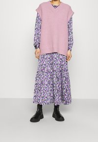 Růžový pletený bezrukoavý svetr s výstřihem do V přes fialové květinové dlouhé šaty. Černé kotníkové boty dotvářejí vrstvený outfit.