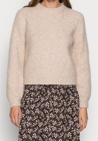 Maglione beige a maglia con maniche lunghe e orlo a coste. Indossato sopra una gonna nera floreale con motivi rosa e giallo. Materiale morbido e testurizzato.