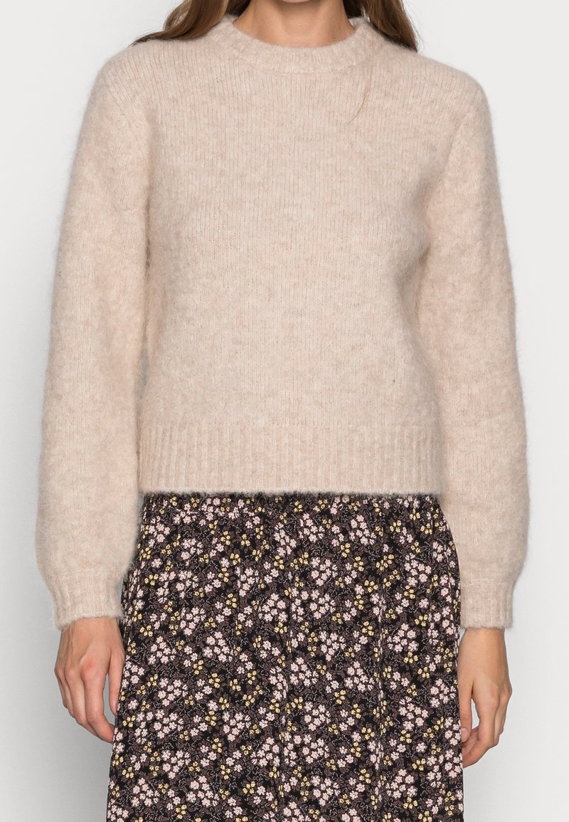 Maglione beige a maglia con maniche lunghe e orlo a coste. Indossato sopra una gonna nera floreale con motivi rosa e giallo. Materiale morbido e testurizzato.