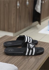 Mustat Adidas-sandaalit, joissa on kolme valkoista raitaa, teksturoitu pohja ja logo hihnassa, aseteltuna puupinnalle pukuhuoneessa.