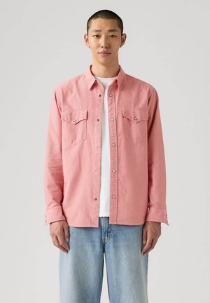 Giovane uomo con capelli corti che indossa una camicia a quadri rossa abbottonata sopra una T-shirt bianca e jeans azzurri, in piedi contro uno sfondo semplice.