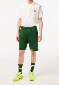 Manlig modell iklädd vit polo t-shirt med logotyp, gröna sportshorts med logotyp, randiga gröna strumpor och neon-gula sneakers som står framför en vit bakgrund.