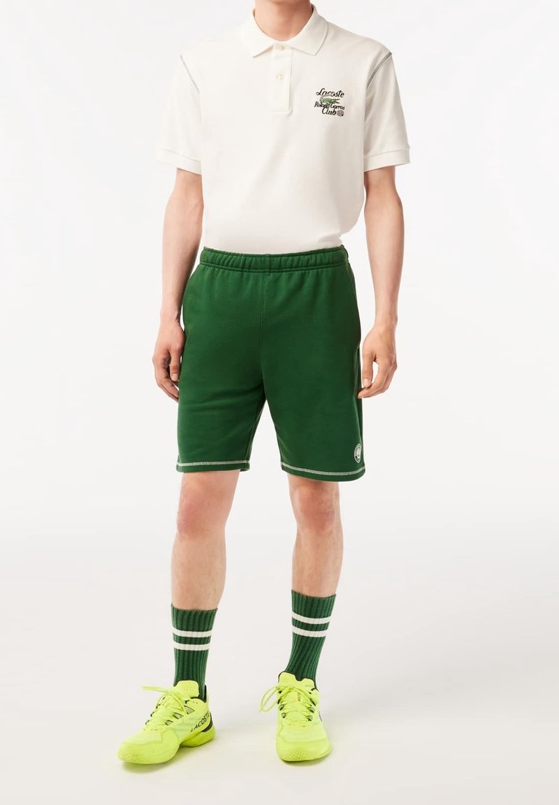 Manlig modell iklädd vit polo t-shirt med logotyp, gröna sportshorts med logotyp, randiga gröna strumpor och neon-gula sneakers som står framför en vit bakgrund.