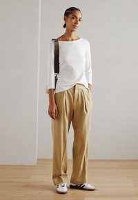 Blusa blanca de manga larga combinada con pantalones plisados beige. La modelo lleva zapatillas blancas con detalles en negro y un bolso de hombro marrón. Fondo de suelo de madera.
