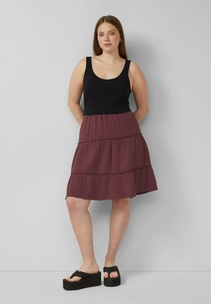 Junge Frau steht, trägt ein schwarzes ärmelloses Top, einen lila gestuften Rock und schwarze Plateausandalen vor einem schlichten hellen Hintergrund.