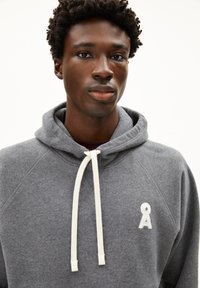 Grijze hoodie met een zachte textuur, voorzien van een voorzak, witte trekkoorden en een klein geborduurd logo op de borst.