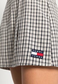 Károvaná sukně z béžové a černé tkaniny s ležérním designem. Obsahuje přišitou nášivku s logem "TOMMY JEANS" v námořnické modré, červené a bílé barvě.