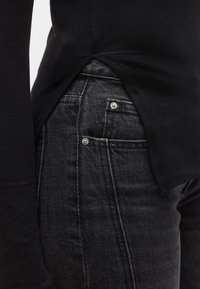 Fekete hosszú ujjú felső magas oldalsliccel, sötétszürke farmernadrággal, amely ezüst színű fémrészletekkel és texturált felülettel rendelkezik.