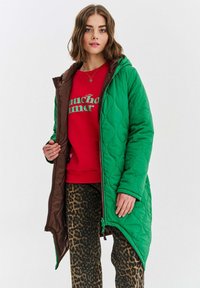 Zelený prešívaný kabát s hnedým interiérom, s kapucňou a zipsom. Nosený cez červenú mikinu a nohavice s leopardím potlačou.