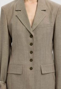 Primer plano de una persona que lleva un blazer topo con cinco botones, solapa de muesca, dos bolsillos con solapa y un bolsillo en el pecho.