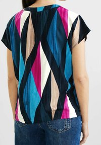 Haut à manches courtes à motifs présentant un mélange de formes géométriques bleues, roses et crème sur un fond sombre, avec une coupe décontractée et un pli au dos.