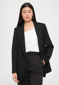 Svart blazer med skreddersydd design, notched lapels og to knapper. Bært over en hvit topp og kombinert med mørke stripete bukser.