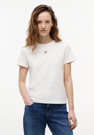 Mujer joven con cabello ondulado, vestida con una camiseta blanca de Tommy Jeans y jeans azules, de pie con una mano en el bolsillo sobre un fondo blanco.