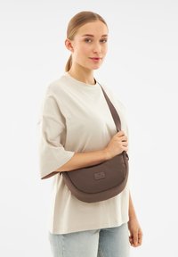 Borsa a tracolla marrone con forma curva, realizzata in materiale morbido, caratterizzata da un patch con logo frontale e una tracolla regolabile.