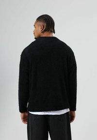 Zwarte, oversized, gestructureerde trui van een pluizige stof. Heeft een ronde halslijn en lange mouwen, gedragen over een wit overhemd.