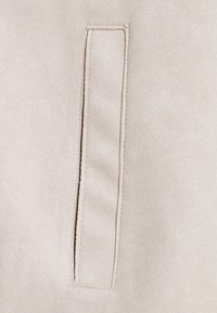 Beige mokkanahka kangas, jossa on pystysuora viillotasku, jossa on ommellut reunat. Kankaassa on pehmeä rakenne ja tasainen, yhtenäinen pinta.