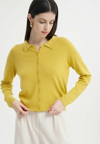 Gelbe gerippte Strickjacke mit kragenfreiem Ausschnitt, Knopfverschluss und langen Ärmeln. Aus weichem, strukturiertem Material mit cropped Passform.