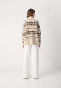 Pull en mailles de couleur beige avec des rayures imprimées marron foncé et crème. Il a un col montant et des poignets côtelés, associé à un pantalon large blanc.