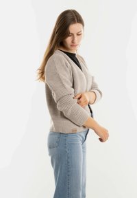 Beige geribde cardigan met driekwartmouwen en een ronde zoom. Gedragen over een zwarte top, gecombineerd met lichtblauwe high-waisted jeans.