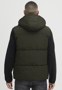 Kapuze Puffer-Weste in dunkelolivgrün mit gestepptem Design, geschichteter Polsterung und einem schwarzen Fleece Langarmshirt darunter.