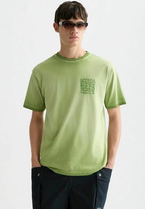 Jonge man met donkere zonnebril, een lichtgroen T-shirt met korte mouwen met kleine tekst op de borst en zwarte broek met zakken.
