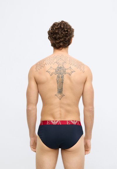 Cuecas azul-marinho com uma cintura elástica vermelha com texto do logotipo, usadas por uma pessoa mostrada de costas, revelando uma grande tatuagem nas costas.