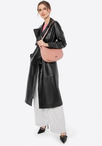 Roze leren handtas met een gladde textuur en ronde vorm, voorzien van een subtiel logo. Model draagt een zwarte leren jas en witte broek.