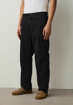 Person trägt schwarze Cargohosen mit großen Seitentaschen, beige Sneaker und ein weißes kurzärmeliges Hemd, steht auf einem grauen Boden.