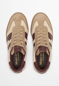 Beige suède sneakers met bordeaux leren strepen, een gestructureerd crèmekleurig paneel, ronde veters en gouden branding op de tong.