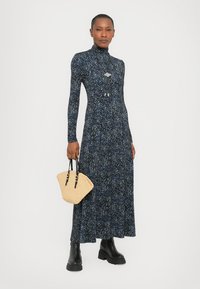 Long-sleeved navy dress with a floral pattern, a high collar, and a flared skirt. Model holds a small woven handbag and wears black boots.