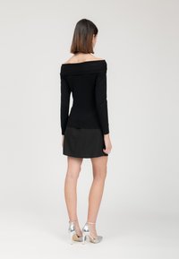 Schwarzes, schulterfreies Langarm-Oberteil, kombiniert mit einem schwarzen Rock, der ein figurnahes Design hat. Das Model trägt silberne High Heels mit Glitzerakzenten.
