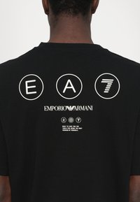 Melna kokvilnas T-kreklā ar baltajiem uzrakstiem: "EA," "7," "Emporio Armani," un tekstuālo saukli; īsām piedurknēm, apaļā kakla izgriezums un gluda tekstūra.