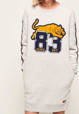 Femme portant une robe sweat-shirt gris clair avec un tigre jaune et un grand numéro 83 bleu marine avec le texte "Super Dry" sur le devant.