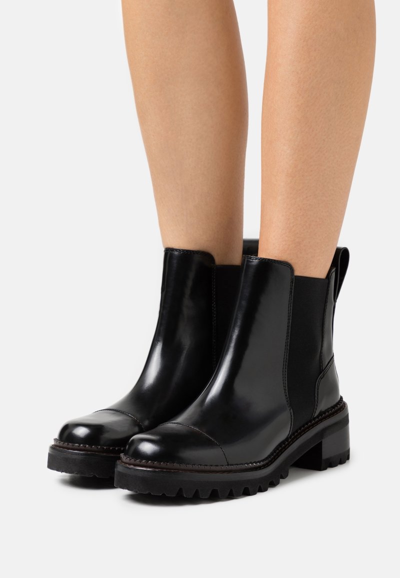 Bottines en cuir noir brillant avec panneaux latéraux élastiques, bouts renforcés, semelles épaisses à crampons, portées sur jambes nues sur fond blanc.