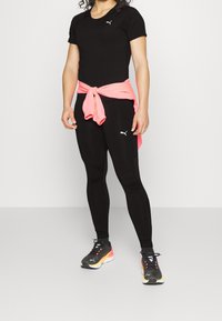 Černé sportovní tričko a legíny v kombinaci s růžovým svázaným svetrem. Černé a barevné běžecké boty s logem.