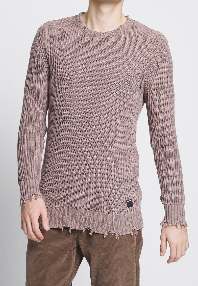 Homme portant un pull en tricot rose poudré avec des bords effilochés et un pantalon en velours côtelé marron, debout devant un fond uni.